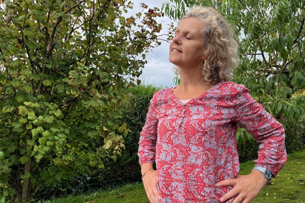 Béa, créatrice de Femmes en Harmonie et femme de 55 ans, se tenant debout dans son jardin verdoyant avec une expression sereine sur son visage. elle symbolise l'importance de penser positivement et de cultiver son bien-être intérieur.