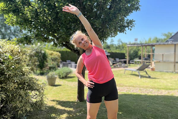 Béa, créatrice de Femmes en Harmonie, souriante et en pleine forme, pratiquant des exercices d'étirement dans son jardin, symbolisant l'accompagnement bienveillant offert à travers le programme '21 jours pour retrouver la forme et l'énergie'.