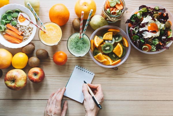 Vue de dessus d'un ensemble d'aliments sains comprenant des fruits, légumes, et smoothies, avec une personne prenant des notes, soulignant l'importance des aliments riches en nutriments pour maintenir un poids équilibré et soutenir le métabolisme après 45 ans.