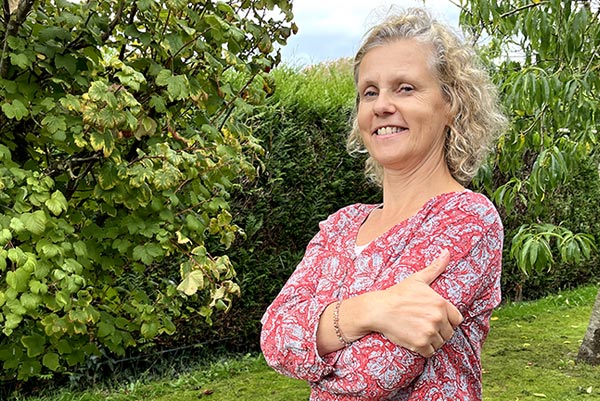 Béa, fondatrice de Femmes en Harmonie, souriante dans son jardin, portant un chemisier fleuri. Un encouragement chaleureux pour aider celles qui le souhaitent à retrouver la motivation et à avancer avec confiance.