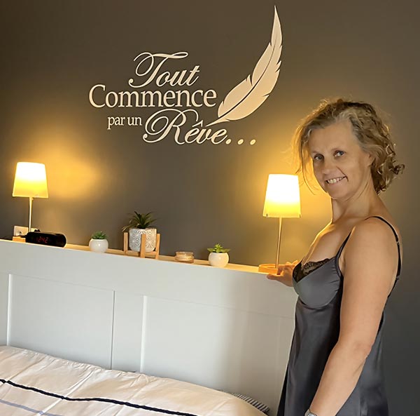 Béa, la fondatrice de Femmes en Harmonie, se tient au pied de son lit, sapprétant à se coucher. Sur le mur, on peut lire l'inscription 'Tout Commence par un Rêve...' 