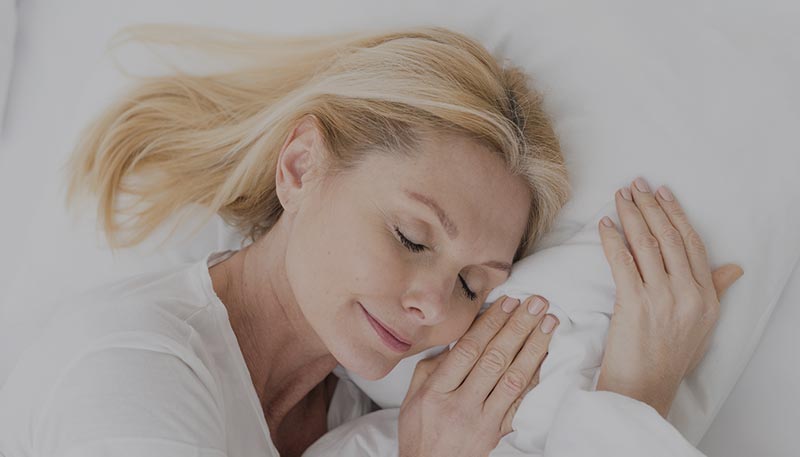 Femme d'environ 50 ans, allongée dans son lit et dormant profondément avec un léger sourire sur les lèvres,. Cette image rappelle l'importance du sommeil pour la santé physique et mentale, Toutes les femmes doivent être sensibles à l'impact d'un bon sommeil sur le bien-être général, la gestion du stress et l'équilibre émotionnel.