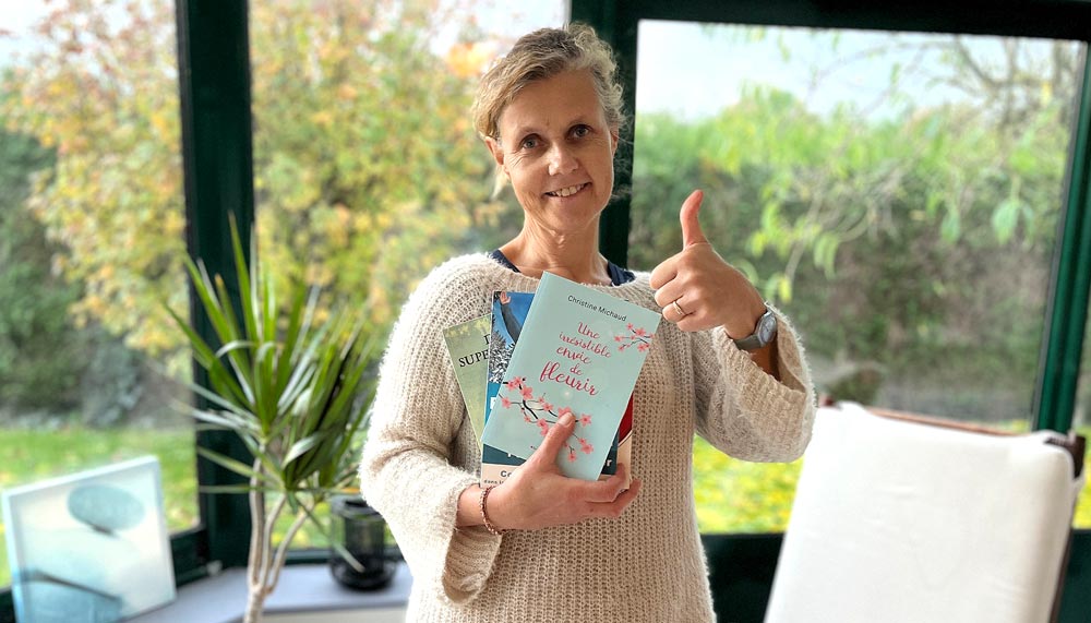 Béa dans sa véranda avec un grand sourire, tenant sous le bras une pile de livre et levant le pouce pour t'encourager à intégrer la lecture dans ton quotidien. 