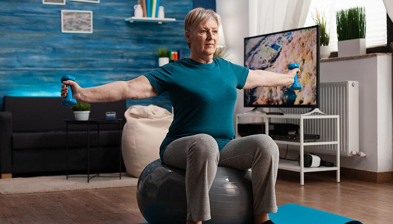 Une femme d'environ 60 ans utilisant des haltères et une balle de fitness à domicile pour améliorer sa posture et renforcer son dos,