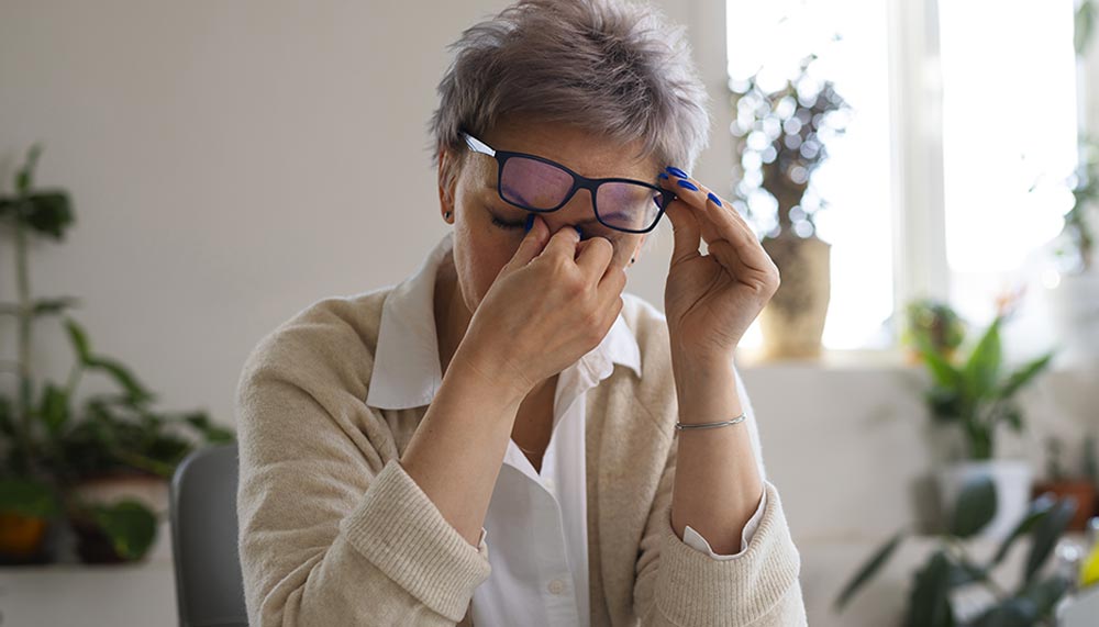 Femme de 55 ou 60 ans épuisée, soulevant ses lunette et se frottant les yeux. La surcharge mentale et le stress chronique sont responsables d’une fatigue émotionnelle intense.